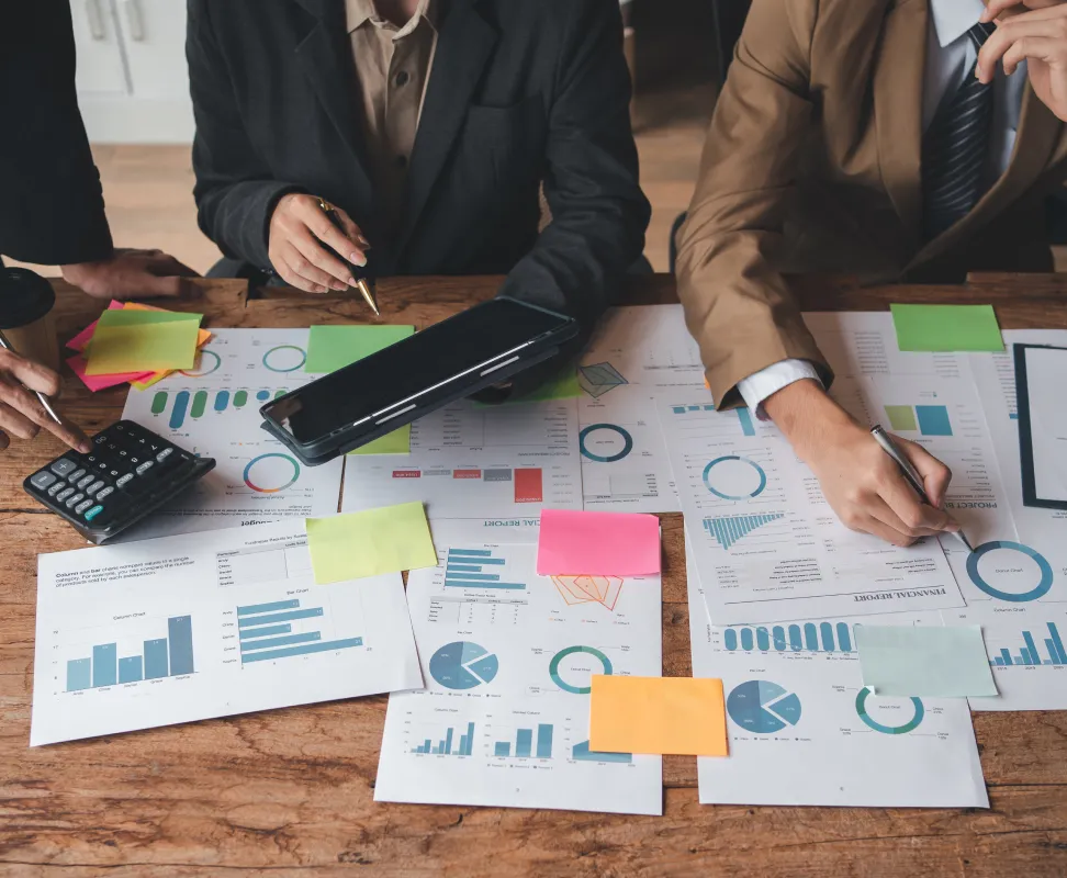 analisis-datos-financieros-negocio Un equipo de negocios colabora sobre una mesa de madera, analizando múltiples informes financieros con gráficos, notas adhesivas de colores y una calculadora para la planificación estratégica.